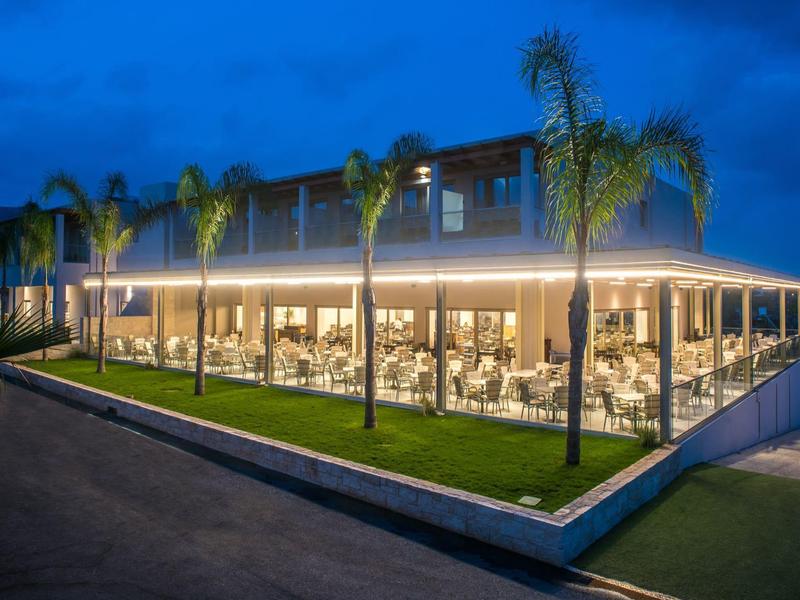 Ristorante illuminato con pareti di vetro e palme all'esterno al crepuscolo.