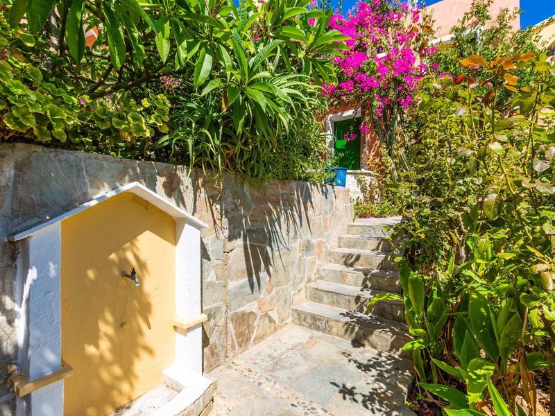 Escalier en pierre à côté des murs de la maison avec une végétation luxuriante et des fleurs roses sous un ciel bleu.