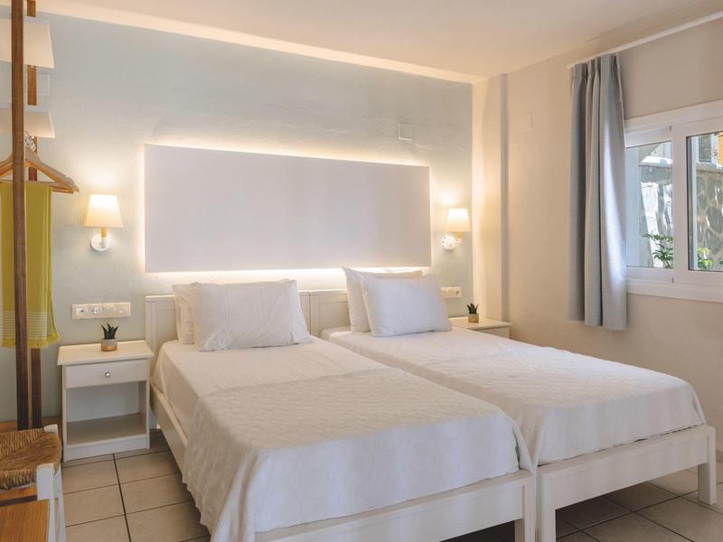 Chambre d'hôtel lumineuse avec deux lits simples, tables de chevet et une fenêtre avec des rideaux gris.