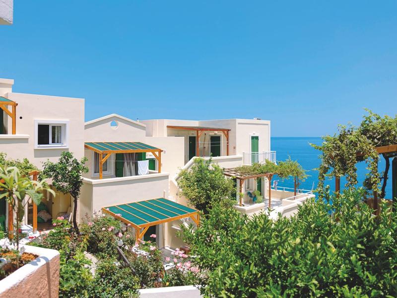 Bâtiments blancs avec des auvents verts entourés de palmiers et vue sur la mer bleue sous un ciel clair.