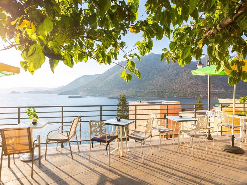 Terrasse ensoleillée avec tables, chaises et vue sur la mer sous des feuilles ombragées.