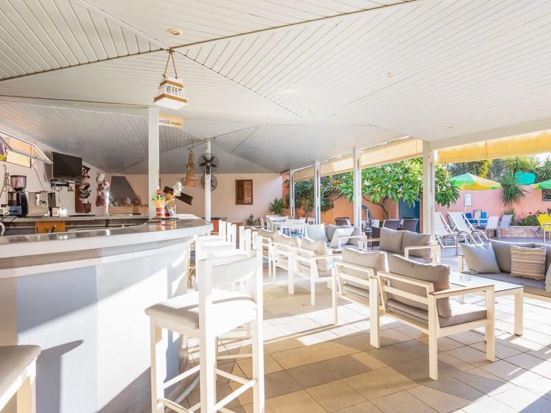 Bar d'hôtel lumineuse avec tabourets blancs et coin salon à côté de grandes fenêtres donnant sur la terrasse.