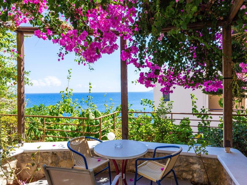 Terrasse avec table et chaises, entourée de fleurs violettes avec vue sur la mer.