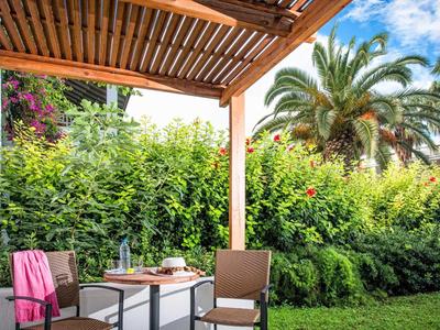 Accogliente patio con tavolo e sedie sotto pergolato in legno in un giardino verdeggiante con palme.