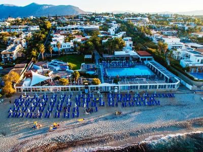 Spiaggia con ombrelloni blu e bianchi e lettini davanti a un complesso di edifici con montagne sullo sfondo.