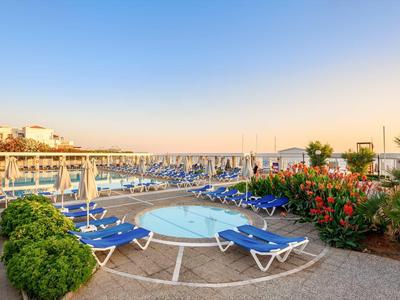 Area piscina dell'hotel con lettini blu, aiuole fiorite e vista sul porto turistico al tramonto.