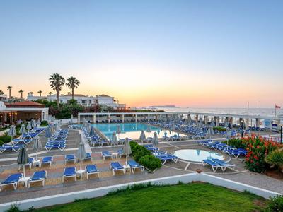 Grande area piscina dell'hotel con molte sedie a sdraio blu e palme al tramonto.