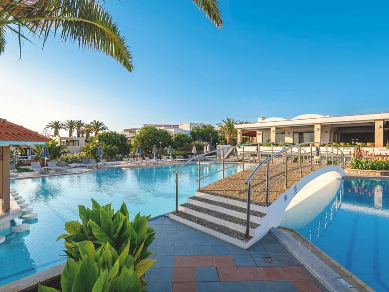 Grande piscina dell'hotel con ponte, circondata da palme e cielo azzurro limpido.