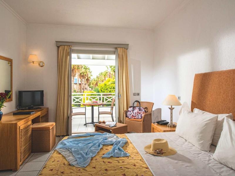 Camera d'hotel luminosa con letto matrimoniale, scrivania, TV, balcone e vista sul giardino.