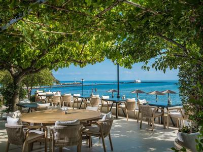Außerhalb sitzendes Restaurant mit runden Tischen unter Bäumen und Blick aufs Meer und Schiffe.