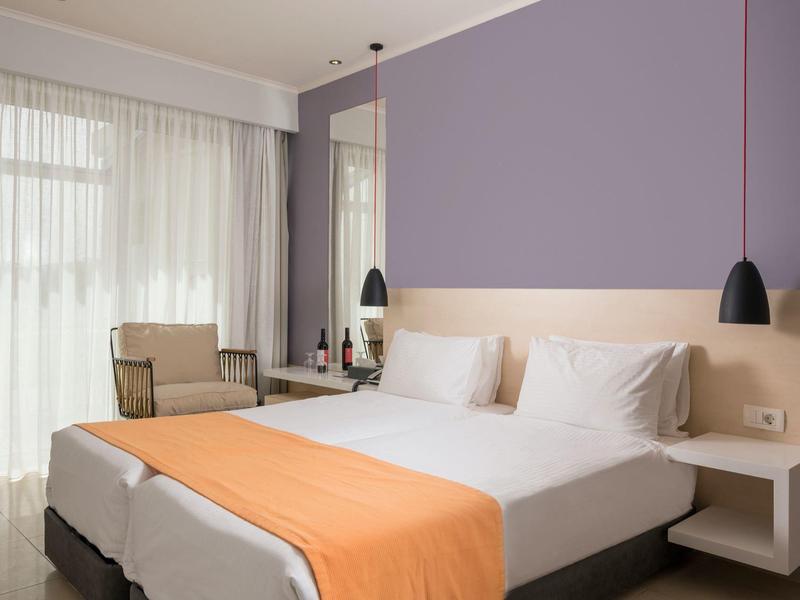 Modernes Hotelzimmer mit großem Bett, oranger Bettdecke und Sitzbereich am Fenster.