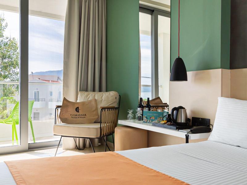 Helles Hotelzimmer mit Bett, grünem Sessel, Balkon und Blick auf Stadt und Berge.