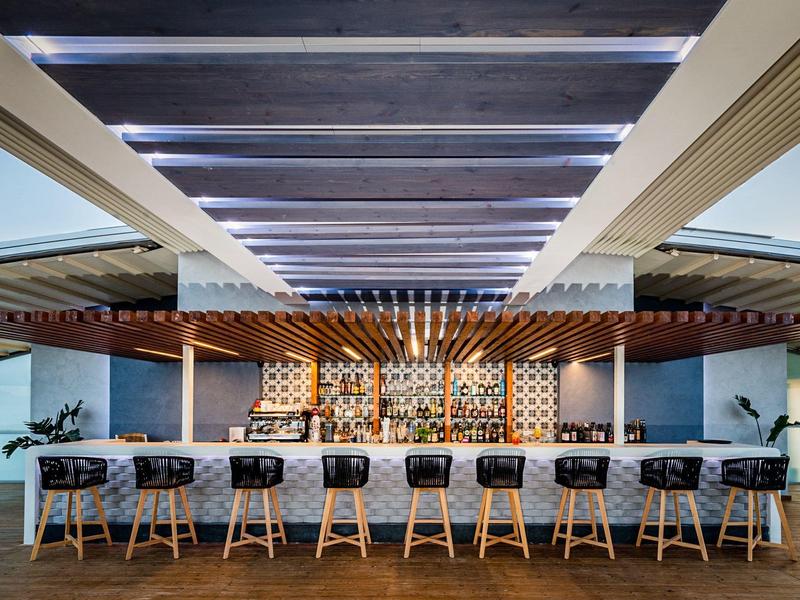 Moderner Hotelbarbereich mit langem weißen Bartresen und sieben Hockern unter einer Holzdecke.