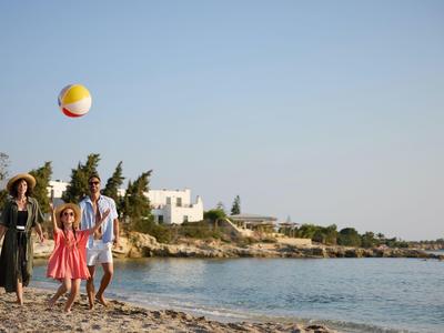 Familie spielt mit einem bunten Strandball am felsigen Meerufer bei klarem Himmel.