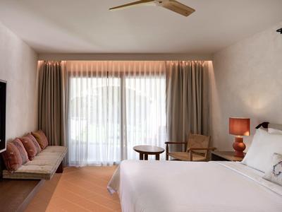 Chambre d'hôtel lumineuse avec grand lit, fauteuils, table et télé murale.