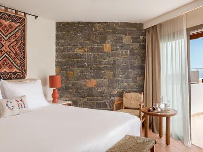 Hell eingerichtetes Hotelzimmer mit großem Bett, Steinglaswand und Zugang zu Balkon mit Meerblick.