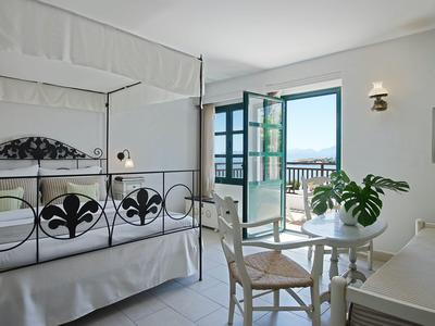 Chambre d'hôtel lumineuse avec lit à baldaquin, table avec chaises et balcon ouvert sur la mer.