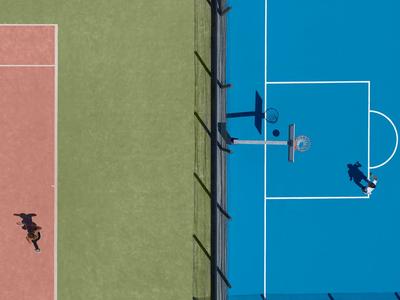 Vista aerea di tre campi sportivi adiacenti in blu, verde e rosso con ombre dei giocatori.