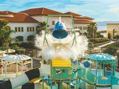 Parc aquatique avec toboggans, grandes fleurs et zone de jeux d'eau devant des bâtiments d'hôtel.
