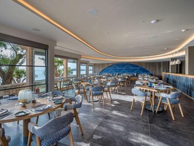 Ristorante moderno con grandi finestre, tavoli in legno e sedie grigie, vista sull'acqua.