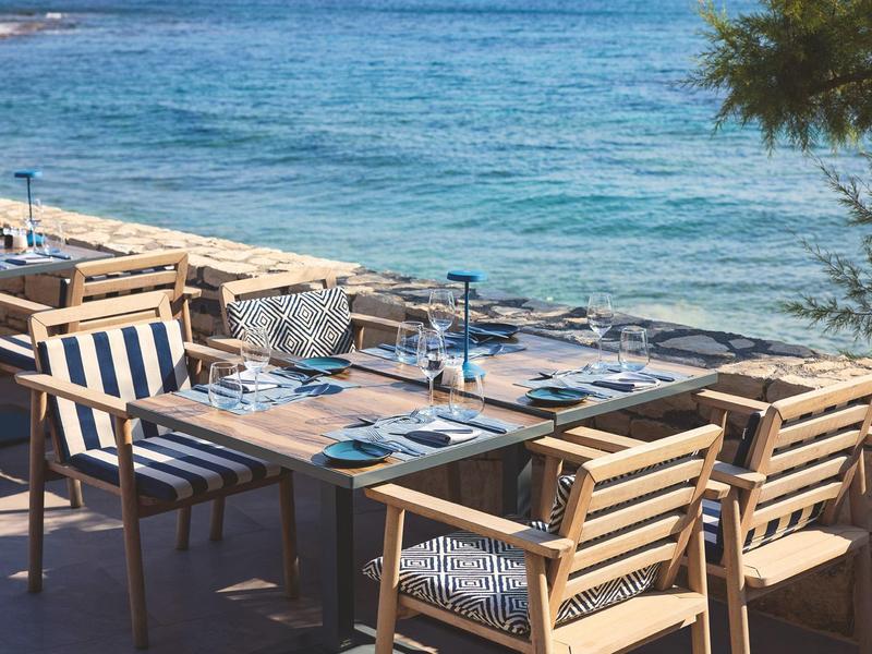 Espace repas avec tables et chaises en bois au bord de la mer avec vue sur l'eau bleue.