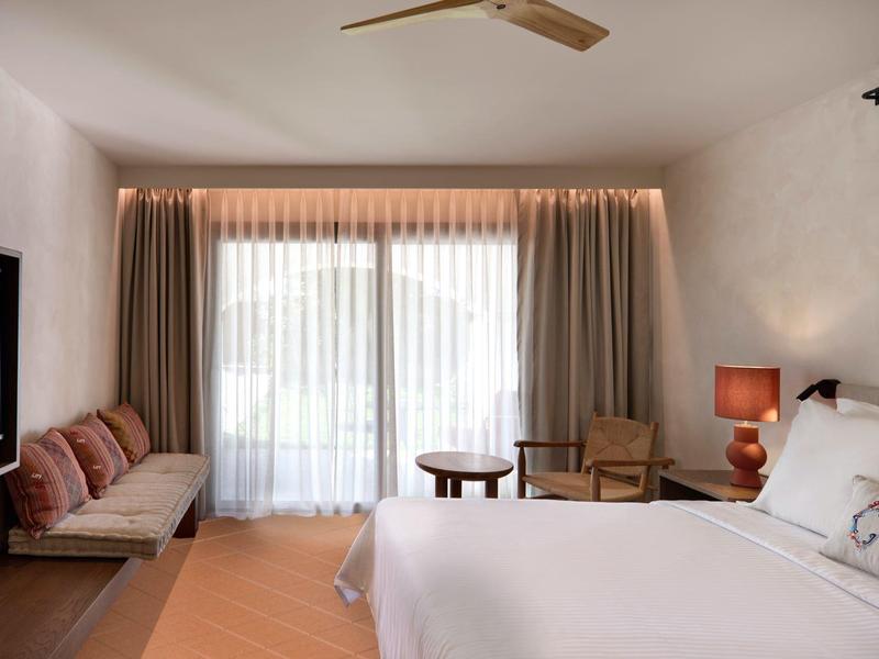 Chambre d'hôtel lumineuse avec grand lit, fauteuils, table et télé murale.