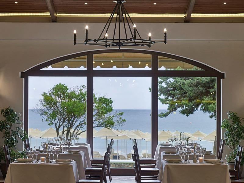 Vue à travers une fenêtre de restaurant sur une plage avec des parasols et des arbres.