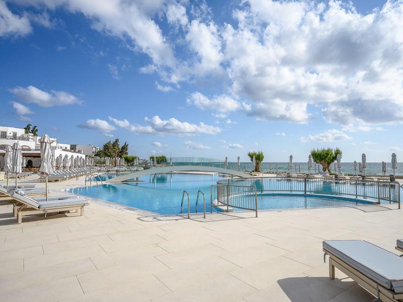 Grand bassin extérieur avec chaises longues, entouré de bâtiments sous un ciel bleu clair.