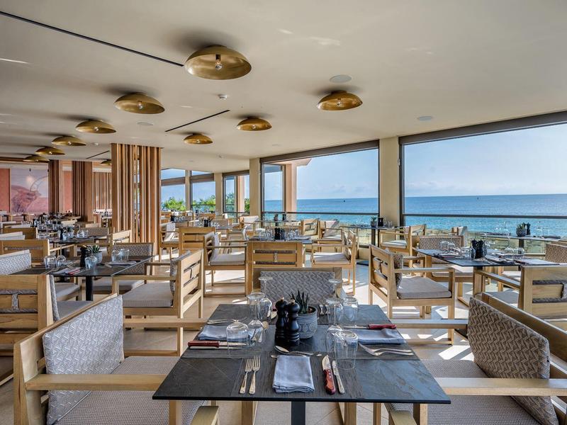 Restaurant moderne avec rangées de tables et vue sur la mer à travers de grandes fenêtres.
