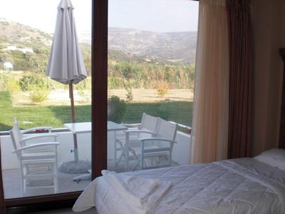 Schlafzimmer mit weißem Bett, Glas-Schiebetür und Terrasse mit Stühlen, Tisch und Sonnenschirm, bergige Landschaft.