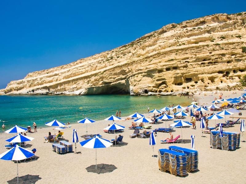 Strand mit blau-weißen Sonnenschirmen, klarem Meer, sandigem Ufer und Felsen im Hintergrund.