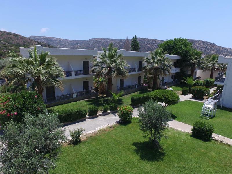 Zweistöckiges Gebäude mit Balkonen, grüner Rasen, Palmen und Gehwege vor bewaldeten Hügeln.