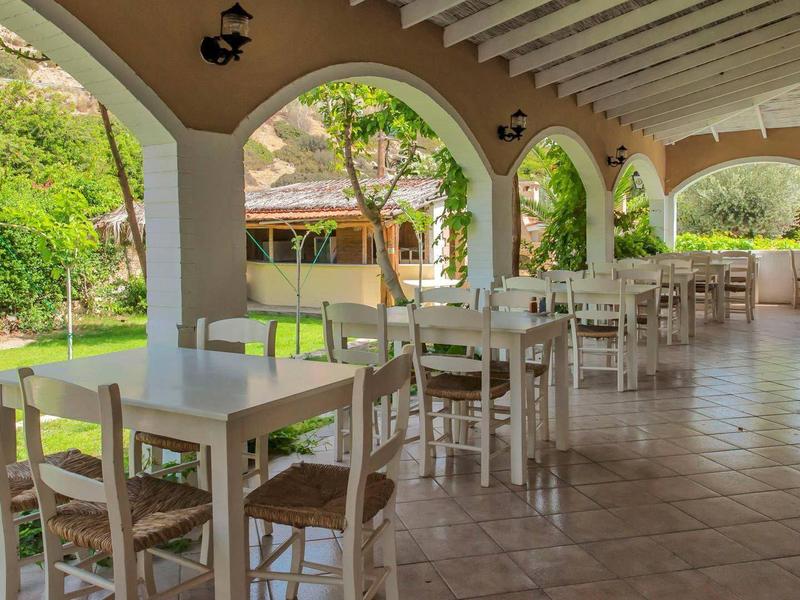 Überdachte Terrasse mit weißen Tischen und Stühlen, offen zu einem grünen Garten mit Hügeln.