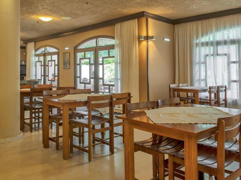 Heller Gastraum mit Holztischen und Stühlen, großen Fenstern und Säulen in warmem Licht.