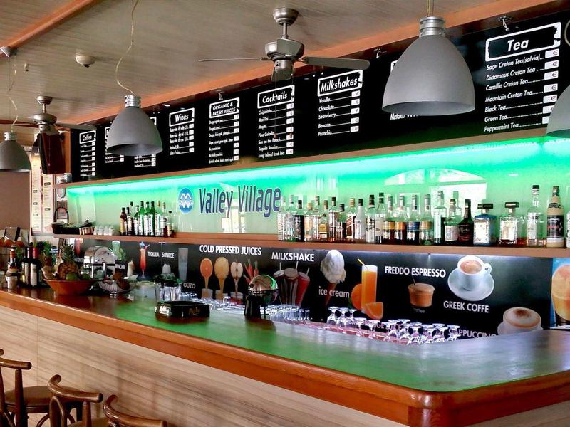 Moderner Barbereich mit Holztheke, grüner Beleuchtung und schwarzer Getränketafel an der Wand.