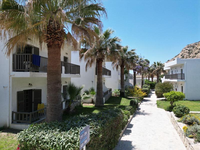 Weiße Häuser mit Balkonen und Palmen entlang eines gepflasterten Weges unter blauem Himmel.