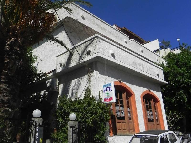 White building with balcony, gate, and small white car under blue sky with palm trees.