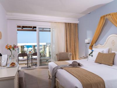 Jasny pokój hotelowy z podwójnym łóżkiem, częścią wypoczynkową i balkonem z widokiem na morze.