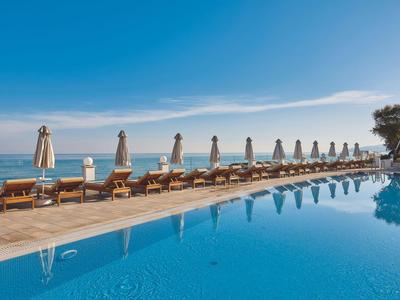 Langer Pool mit Sonnenschirmen und Liegestühlen am Meer unter blauem Himmel.