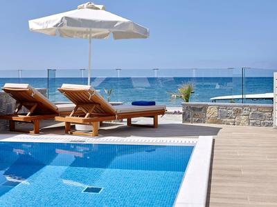 Poolbereich mit zwei Liegestühlen, Sonnenschirm und Meerblick bei klarem Himmel.