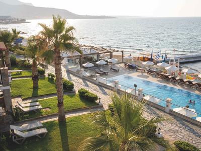 Hotelpool mit Liegestühlen am Meer und Palmen im Gartenbereich bei Sonnenlicht.