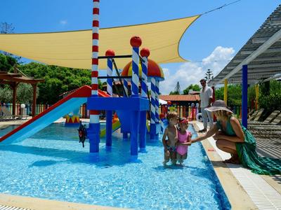 Dzieci bawiące się w basenie z kolorowym placem zabaw wodnym na zewnątrz hotelu w słoneczny dzień.