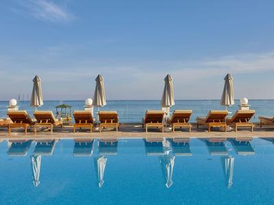 Řada lehátek s deštníky u bazénu s výhledem na moře pod modrou oblohou.