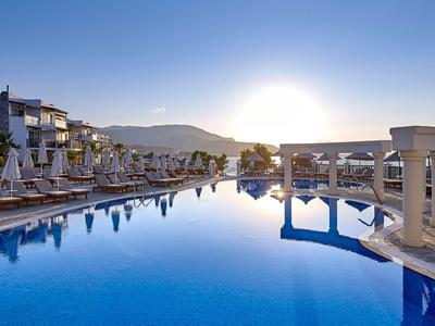 Velký hotelový bazén s lehátky při západu slunce pod jasnou modrou oblohou.