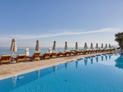 Dlouhý bazén s lehátky a slunečníky u moře pod jasnou oblohou.