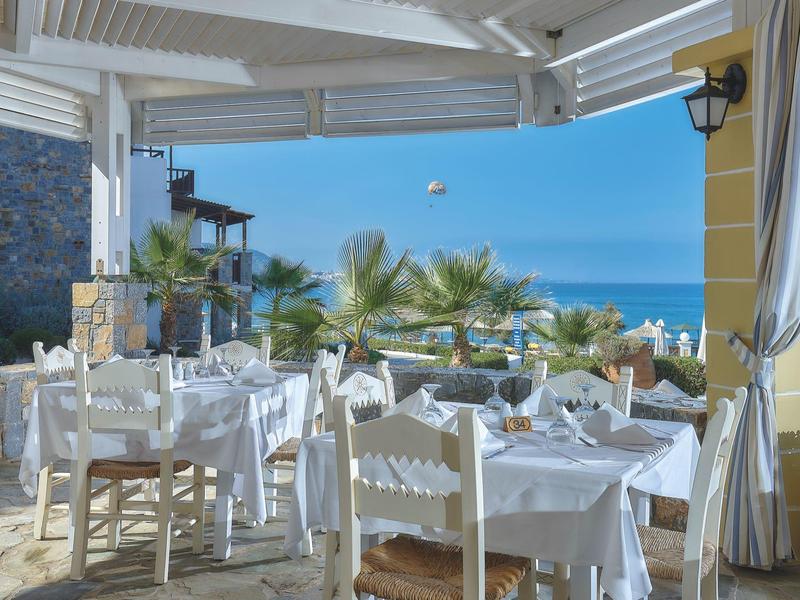 Restauracja na świeżym powietrzu z białymi stołami i krzesłami, widokiem na morze i palmy w tle.