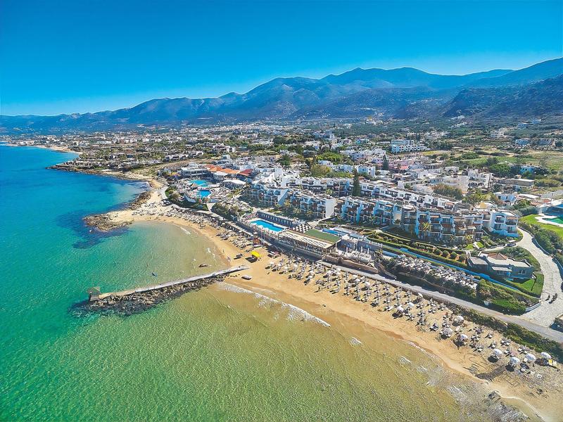 Widok z lotu ptaka na plażę z czystą wodą, opalających się, hotelami i górami w tle.