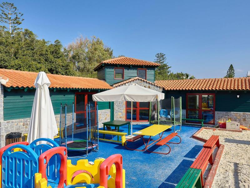 Plac zabaw dla dzieci z huśtawkami, piaskownicą i kolorowymi zabawkami obok budynku z czerwonym dachem.