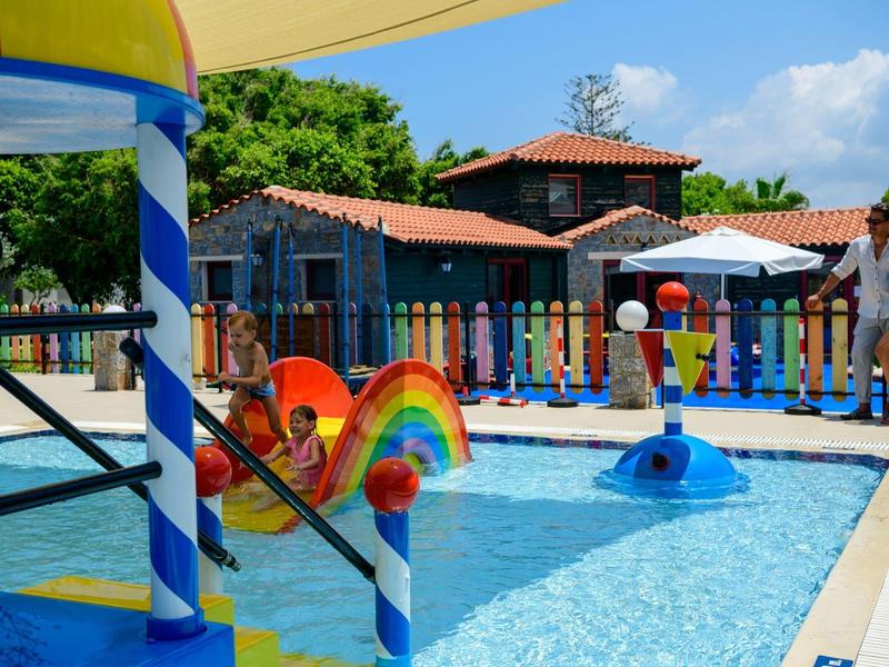 Brodzik dla dzieci z tęczową zjeżdżalnią i daszkiem przeciwsłonecznym obok budynków hotelowych w słoneczny dzień.