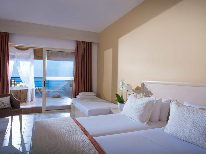 Chambre d'hôtel lumineuse avec deux lits, chaise, balcon et vue sur la mer.